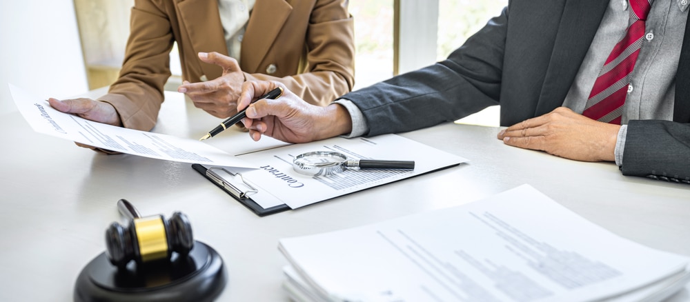Two people in business attire review a contract at a desk with a gavel, magnifying glass, and documents. One points while the other holds a pen over the contract.